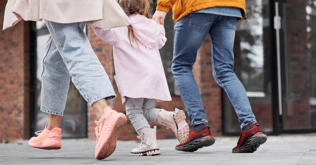 A mother and a father hold a little girl's hands as they walk down a city street.