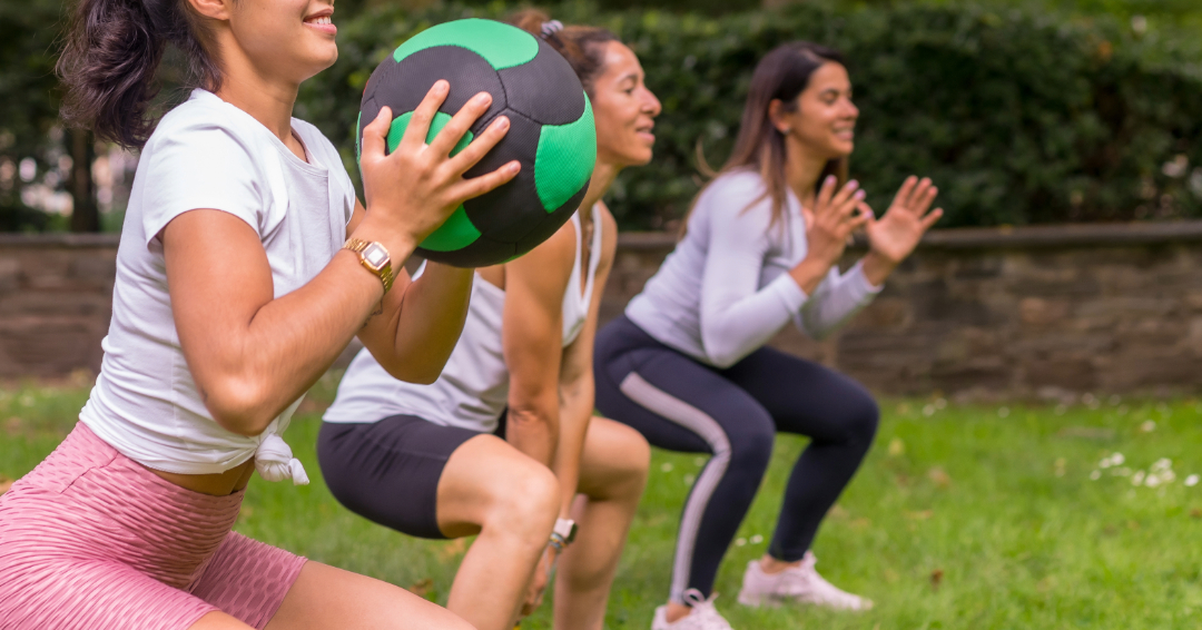 Three women are doing squats with medicine balls in an outdoor setting. 