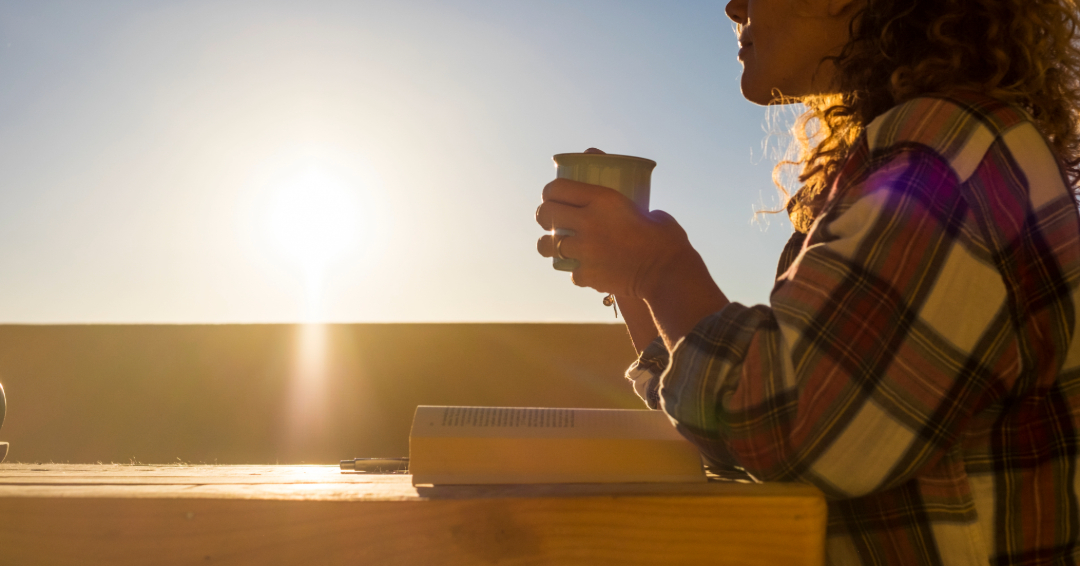A woman sits with an open book and a cup of coffee, gazing over a sunrise. 