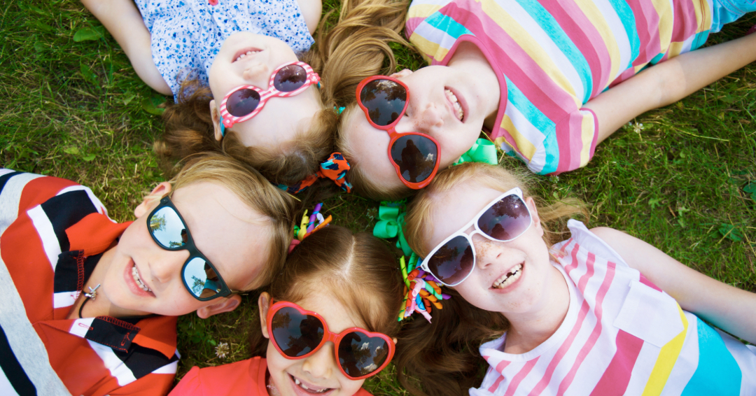 A circle of kids lays on the grass, all wearing colorful sunglasses. 
