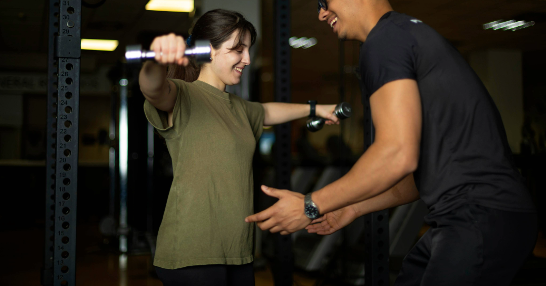 A personal trainer leads a woman in an arm exercise, where she extends weights away from her body.