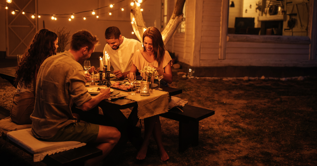 Four people laugh around a table, having an outdoor dinner under string lights. 