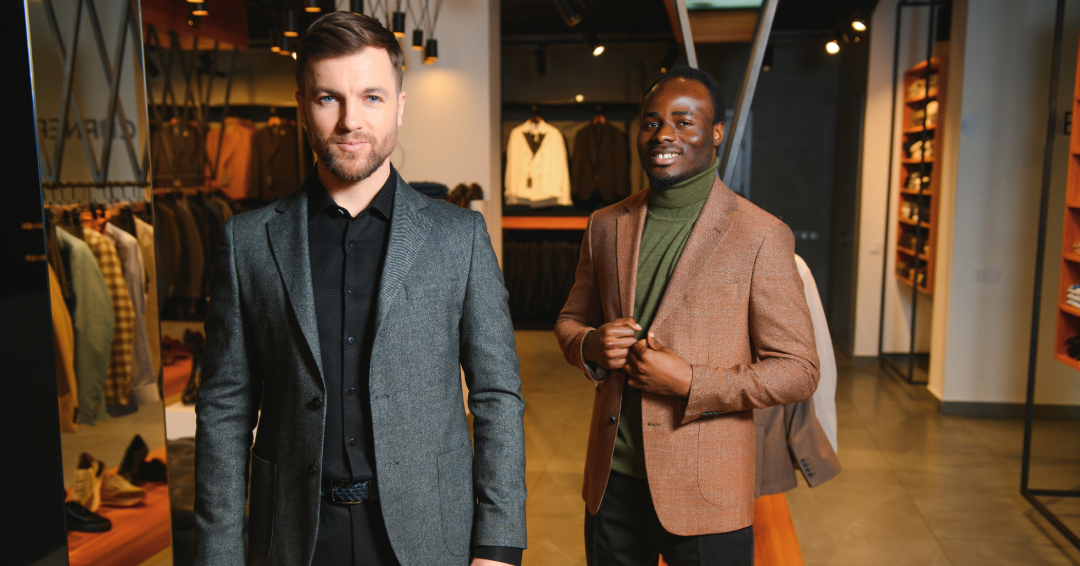 Two well-dressed men pose inside a modern menswear store, one in a gray blazer over black and the other in a brown blazer with a green turtleneck, showcasing classic and contemporary fashion. 